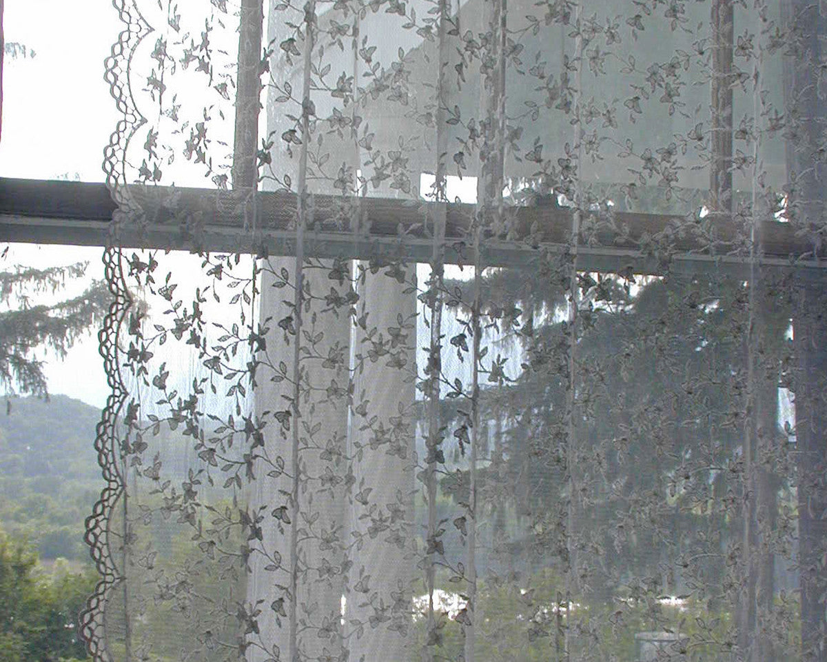 A close-up view of a window adorned with a delicate sheer netting panel featuring butterfly-like decorations. Soft folds and scalloped sides add a charming touch. Available in lengths of 84 inches and 96 inches.